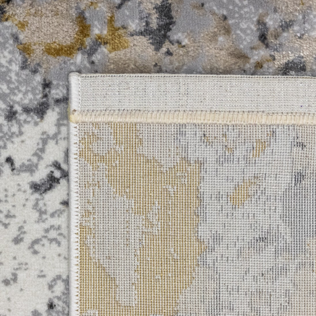 Paradise Living – Tapete Batik 38/017 Cinzento, detalhe do canto com verso resistente ao desgaste e costura de rebordo dupla, padrão cinzento e dourado visível