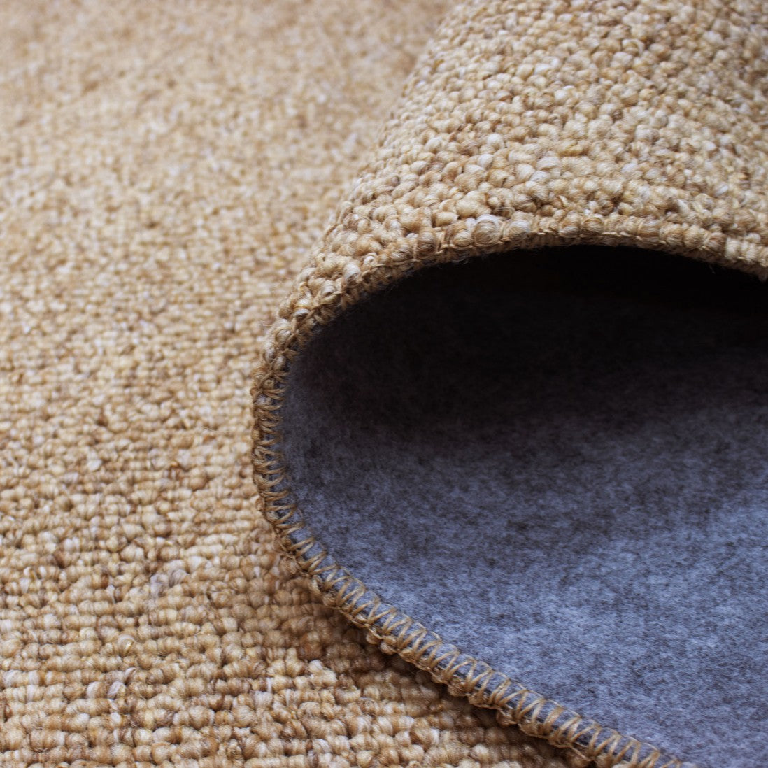 Paradise Living – Tapete Polo 71511/050 Bege close-up do canto enrolado mostrando textura de lã bouclé bege e verso em feltro cinzento antiderrapante