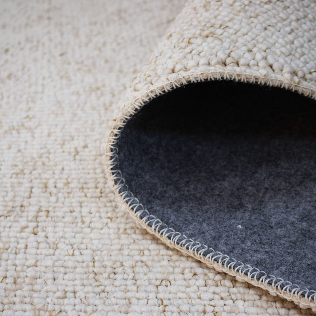 Paradise Living – Tapete Polo 71511/056 Bege Creme close-up do canto enrolado mostrando textura de lã bouclé creme e verso em feltro cinzento antiderrapante