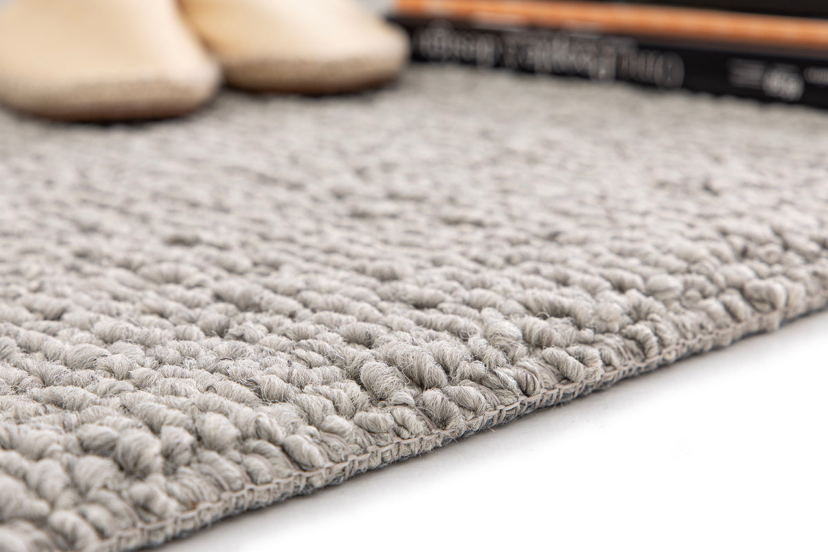 Paradise Living – Tapete Bhutan 71591/060 Cinzento close-up do canto com textura bouclé volumosa em cinzento e chinelos bege ao fundo