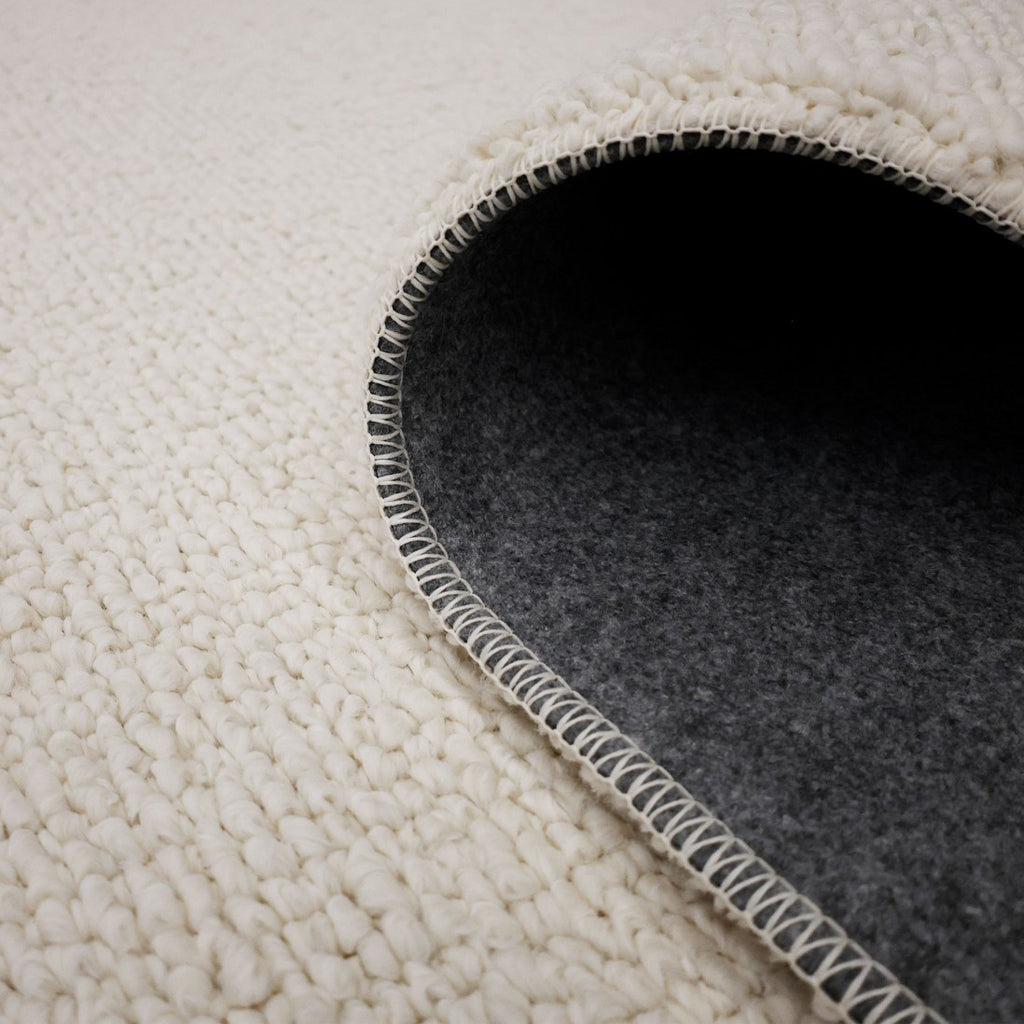 Paradise Living – Tapete Bhutan 71591/066 Branco close-up do canto enrolado mostrando textura bouclé branca e verso em feltro cinzento antiderrapante